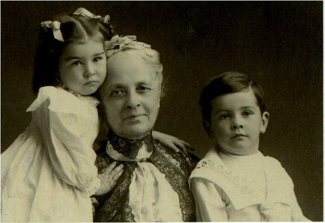Woman with young girl wearing white dress and ribbons in hair.  Young boy wearing white suit.