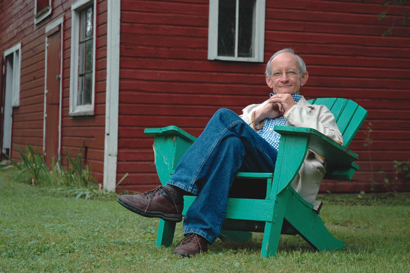 Ted Kooser sits in a green adirondack chair outside a red and white barn.
