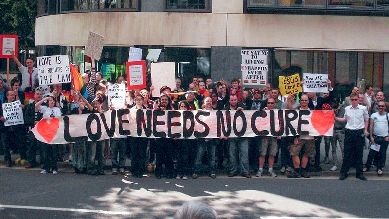 LGBTQ+ protesters hold a banner that says 'Love needs no cure'; links to news story