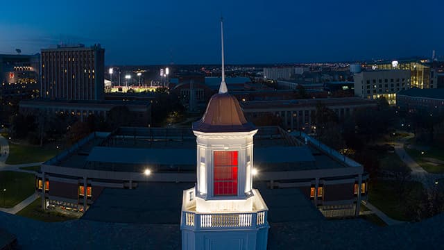 Love Library cupola is illuminated with red light at sunset with campus skyline in background.
