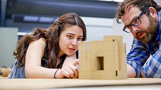 A student and professor discuss the structural openings and angles on an architectural model on a desk.