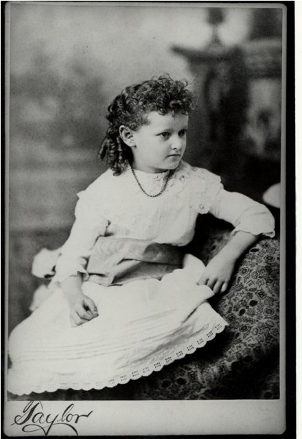 Young girl approximately 5 years only wearing a dress and necklace leaning against a couch. Photographer is Taylor.