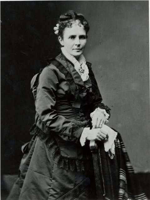 Woman in long dress holding a handkerchief while standing behind a chair.