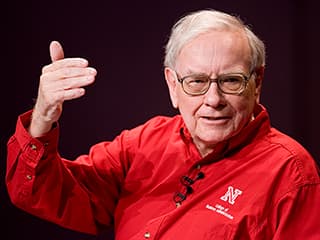 Warren Buffet, wearing a red long sleeve Nebraska shirt, raises his right hand to illustrate a point while talking.