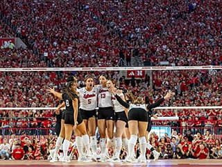 The Husker Volleyball team huddles at the net with the record-setting crowd behind them at Memorial Stadium.