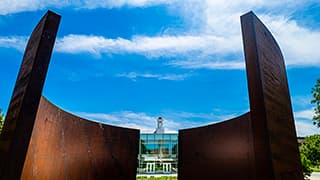 Richard Serra's Greenpoint sculpture frames the Love Library cupola under bright blue skies and wispy clouds on City Campus.