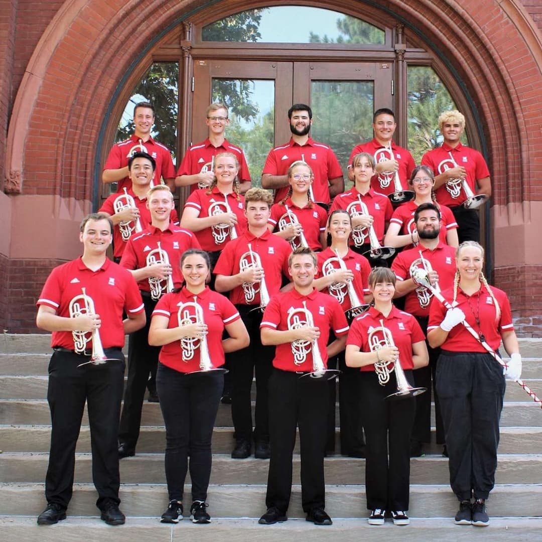A group of brass players in red polos and black pants pose with their instruments on the steps in front of Architecture Hall.