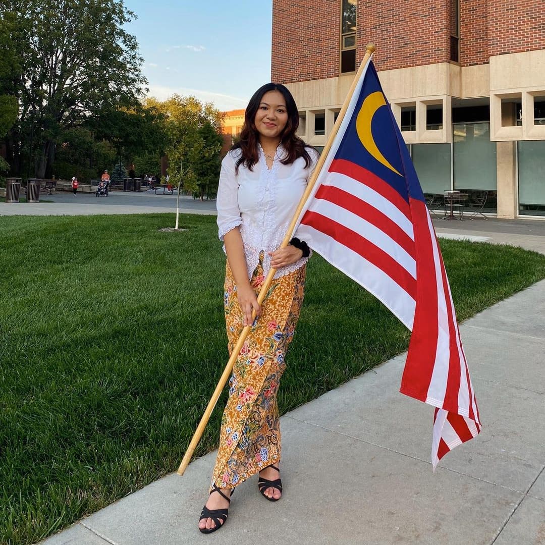 A student smiles and holds a Malaysian flag on a flagpole.