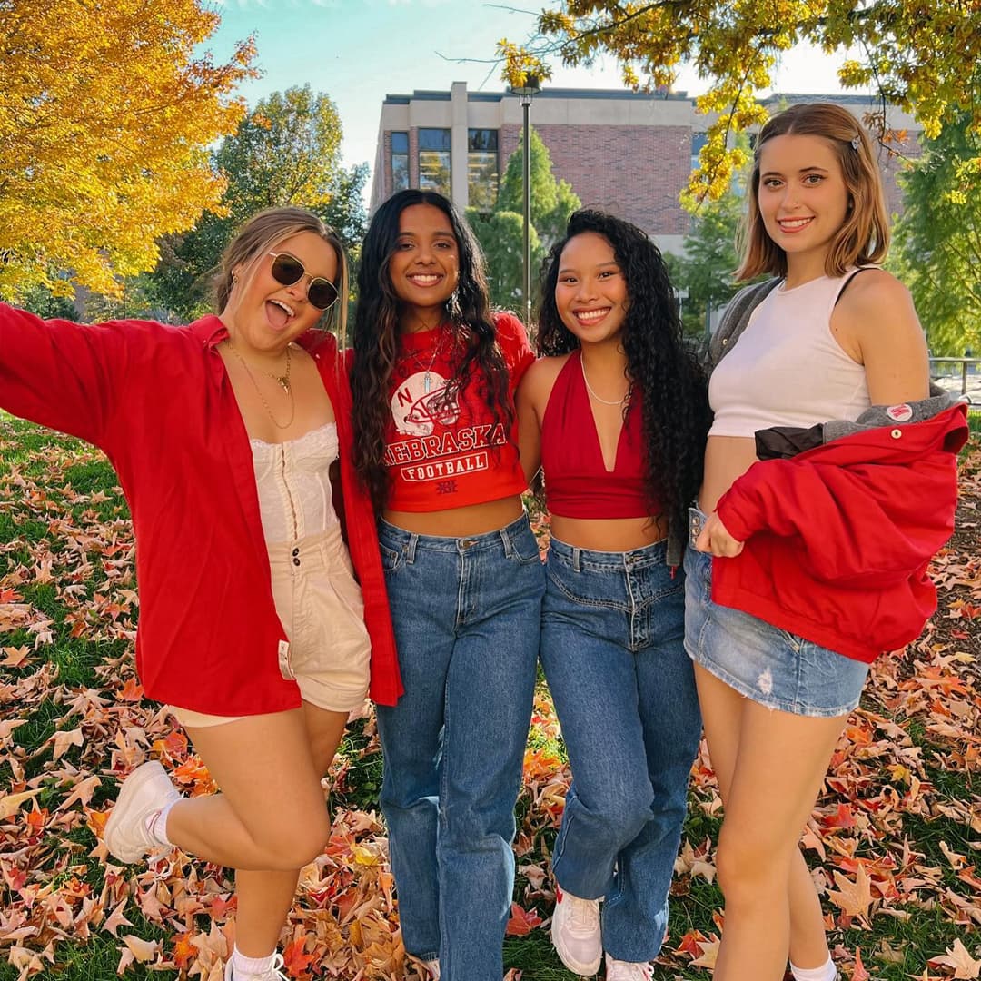 Four Huskers pose for a pic with fall leaves on the trees and ground.
