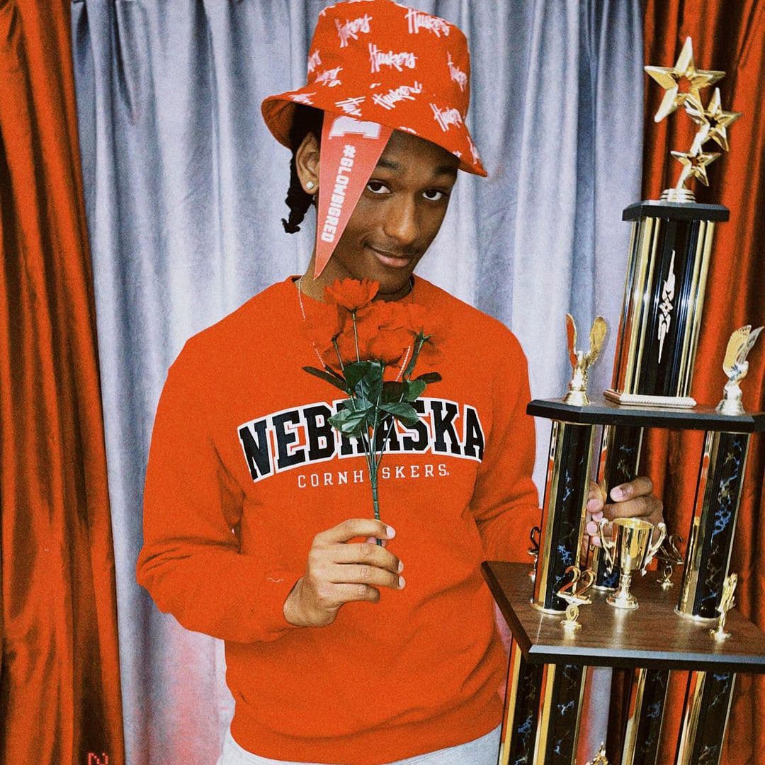 A student wearing a red Nebraska Cornhuskers sweatshirt and Huskers bucket hat holds a red rose in one hand and a trophy in the other.