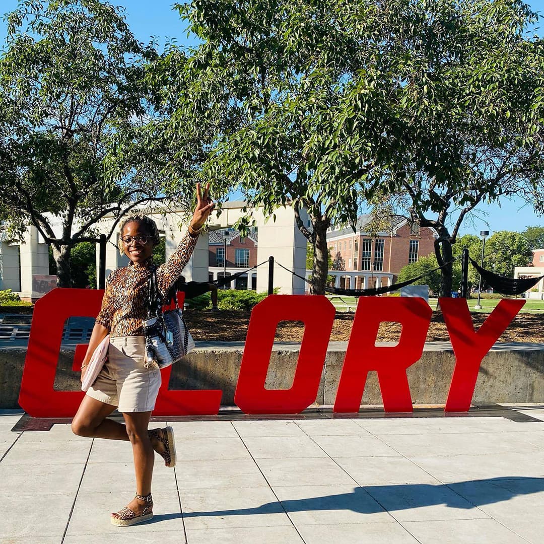Smiling student raises arm and two fingers to make peace sign in front of a large, metal sign that reads GLORY.