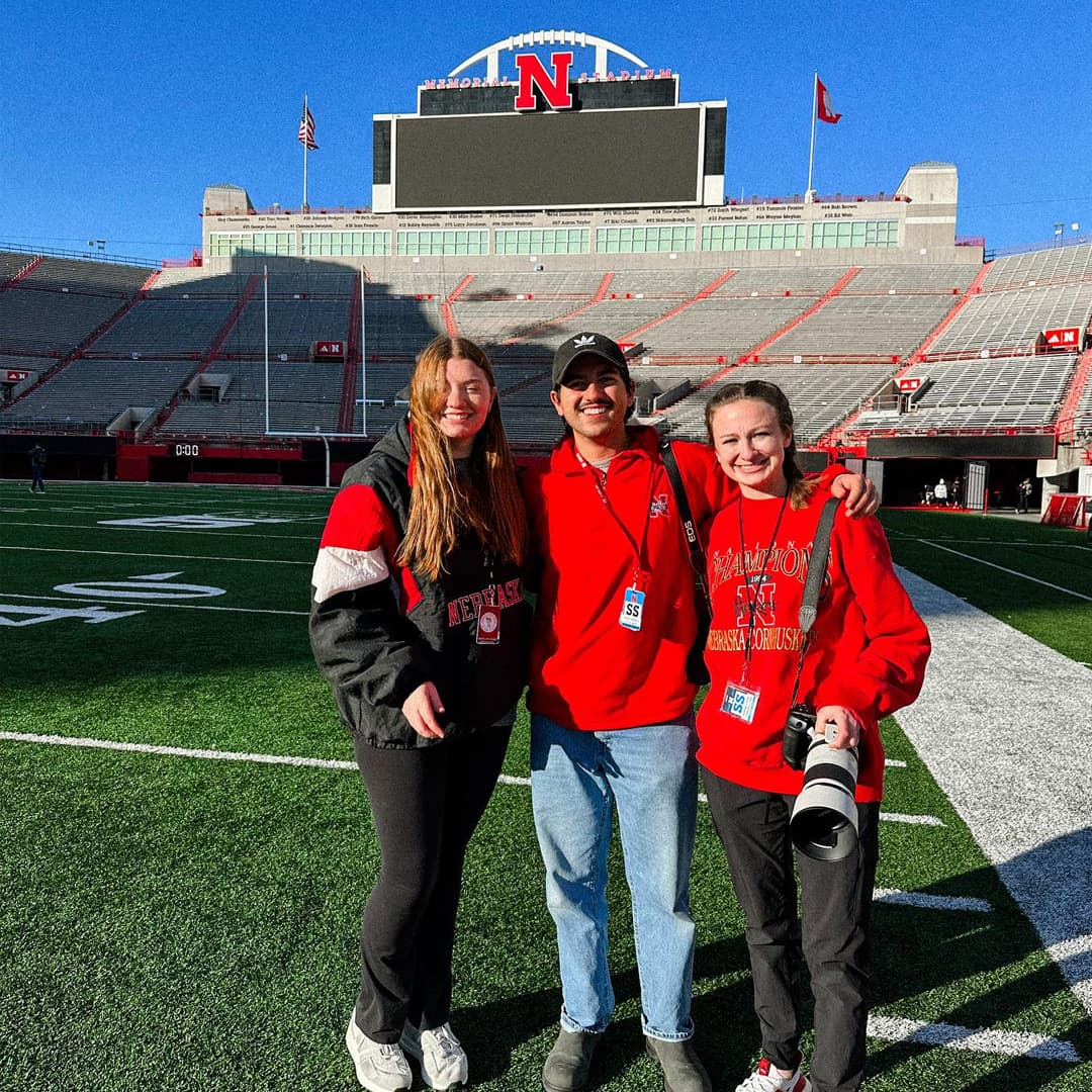 Three Huskers (one holding a camera) smile while on the field at an empty Memorial Stadium on a sunny day.