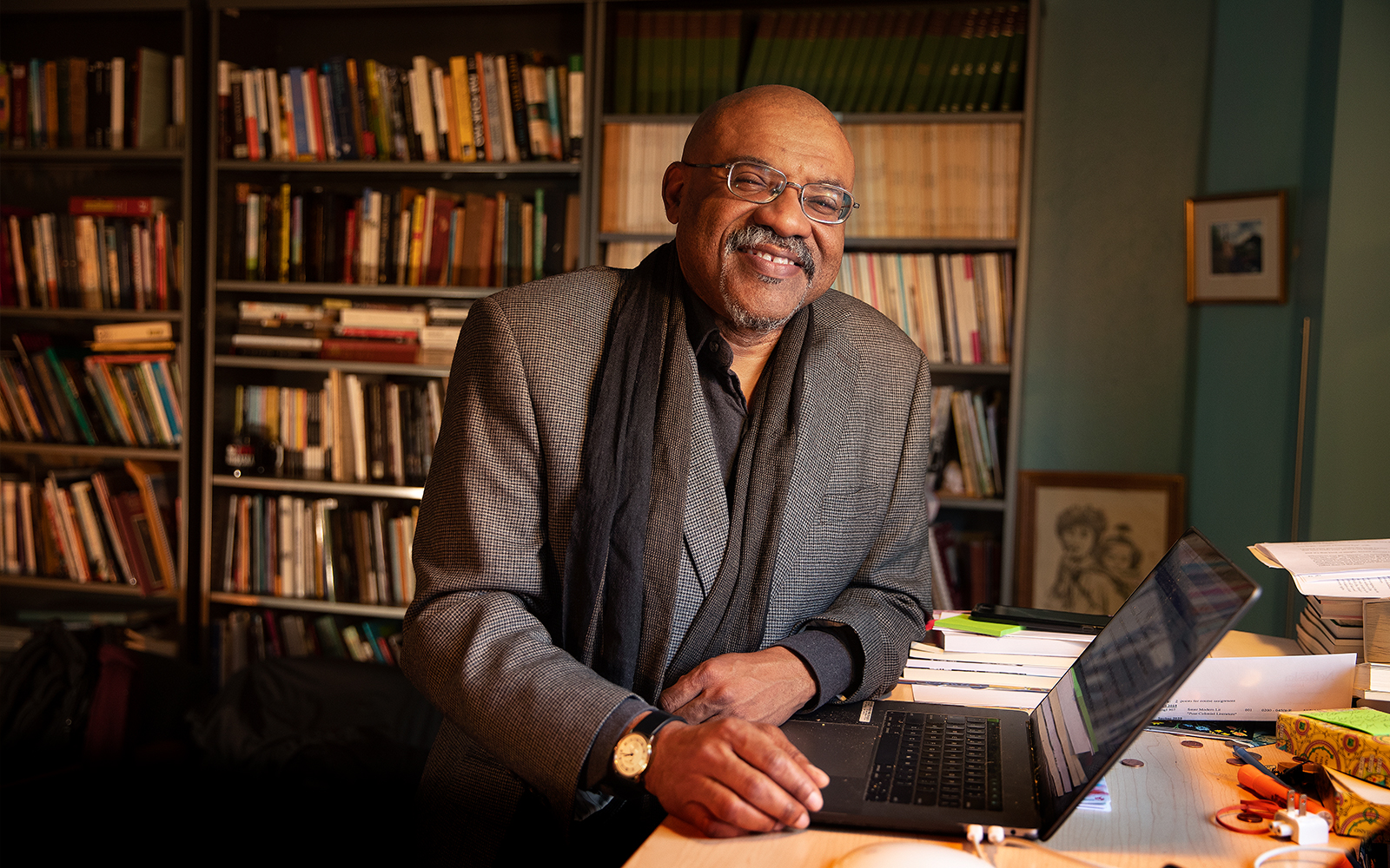 Kwame Dawes stands at his desk with books and past publications of Prairie Schooner behind him