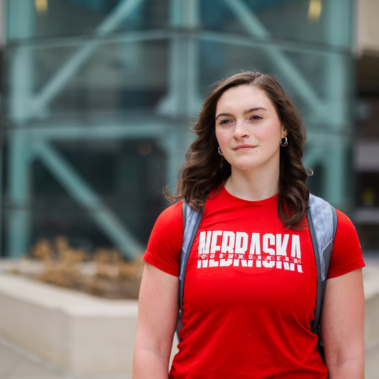 Jozy Piper in a red Nebraska shirt with a backpack, standing outdoors near modern glass architecture.