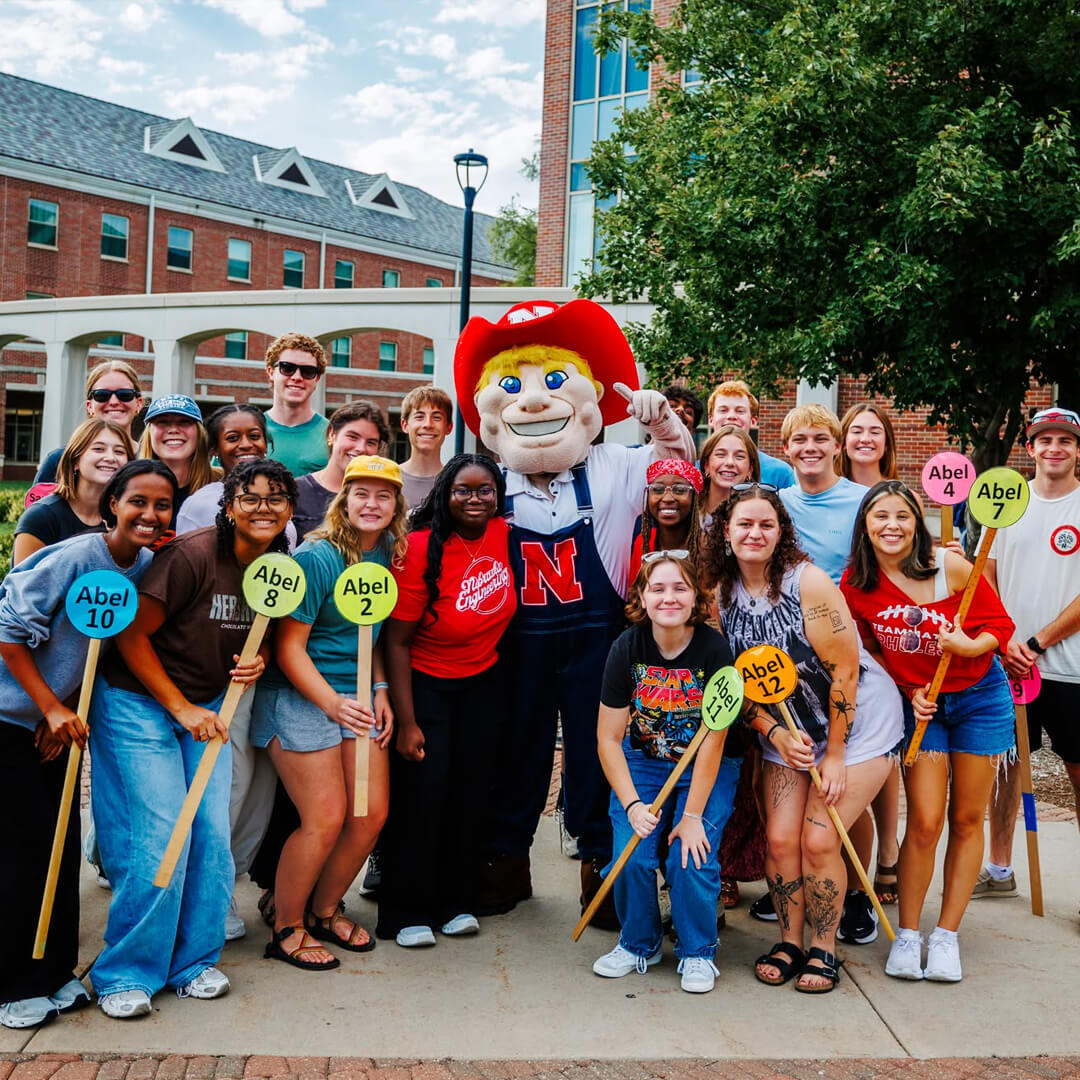 Group of students smiling outdoors with Herbie Husker in red hat, holding colorful signs near a brick building.
