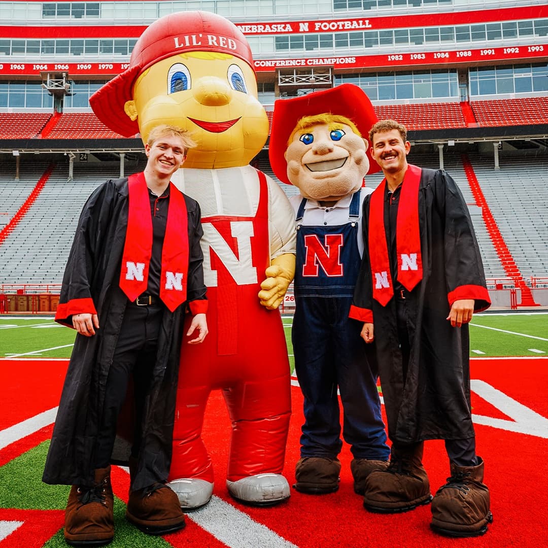 2026 graduating mascots Aidan Kauth-Fisher and Connor Hadaway pose with Lil’ Red and Herbie Husker (respectively) on the field at Memoiral Stadium.