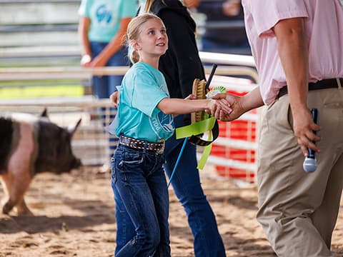 Young girl shaking hands at a livestock show, holding a ribbon, with a pig in the background.