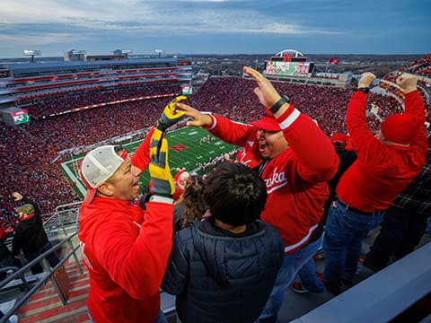 Fans celebrate in a packed football stadium, wearing red, with the game visible under a cloudy sky.