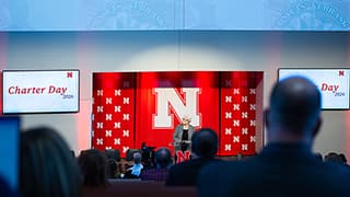 Katherine Ankerson, interim University of Nebraska–Lincoln chancellor, stands at a podium and speaks during the university's Charter Day celebration on February 13. A white Nebraska N on a red background is behind her. The backs of audience members' heads are in the foreground.