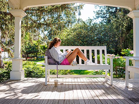 Woman relaxing on a white bench, reading in a sunny gazebo surrounded by greenery.