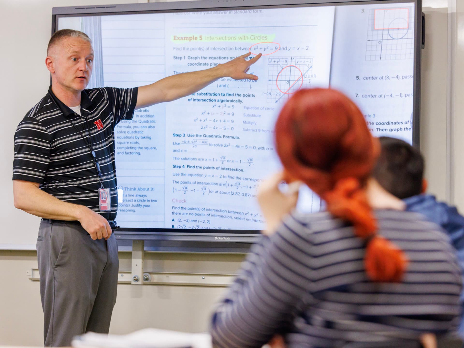Teacher pointing to math equations on a screen while students listen attentively.