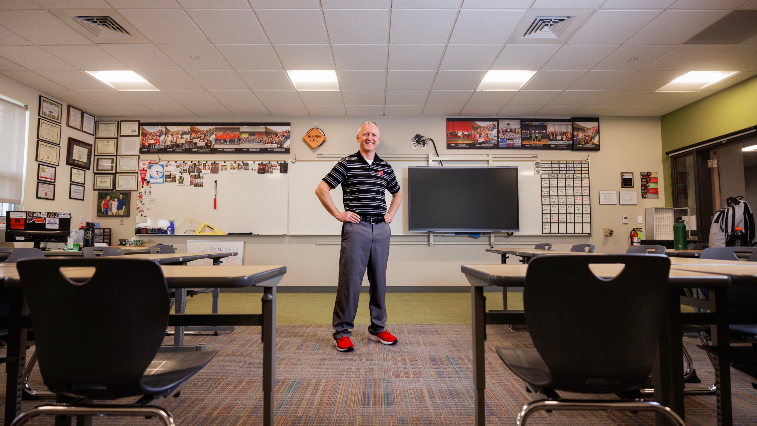 Classroom with a man standing confidently, surrounded by desks and educational decor.