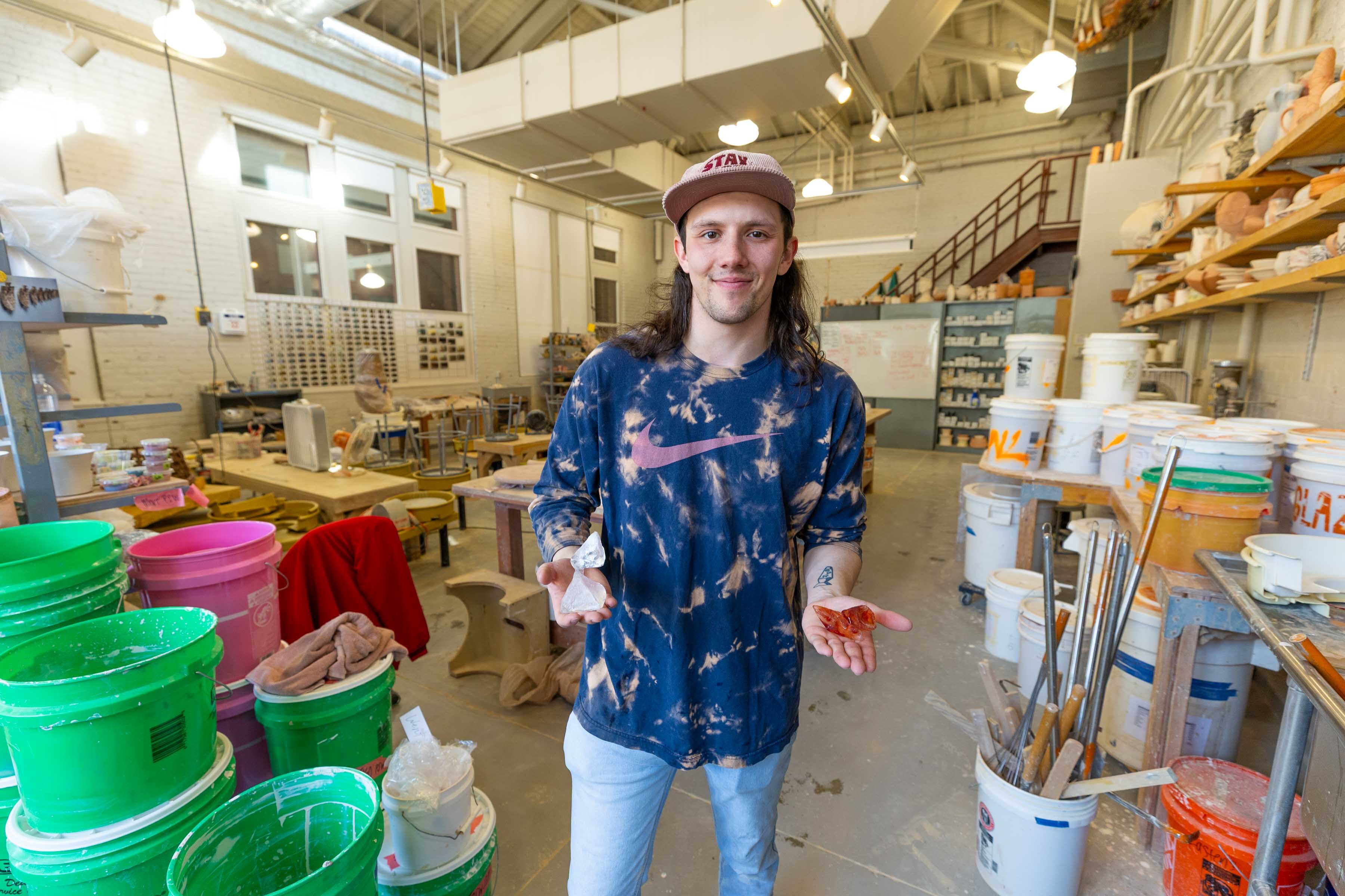Levi Robertson in a colorful art studio in Richards Hall holding small glass sculptures made for his senior capstone, surrounded by buckets, tools, and shelves.