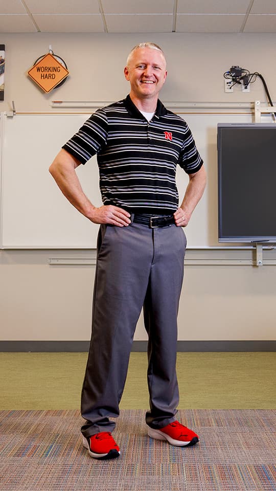 Smiling man in a striped shirt and red shoes, standing in a classroom near a whiteboard.
