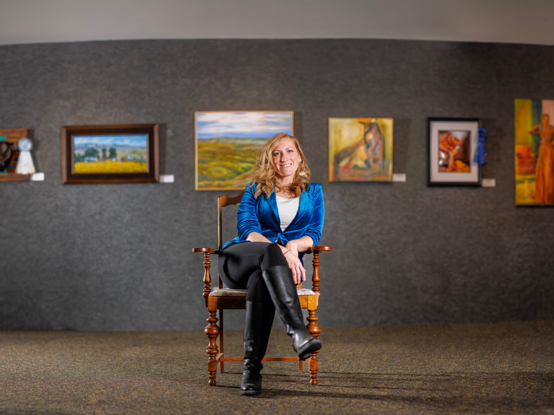 Woman seated on a wooden chair, smiling, in an art gallery with colorful paintings on the wall.