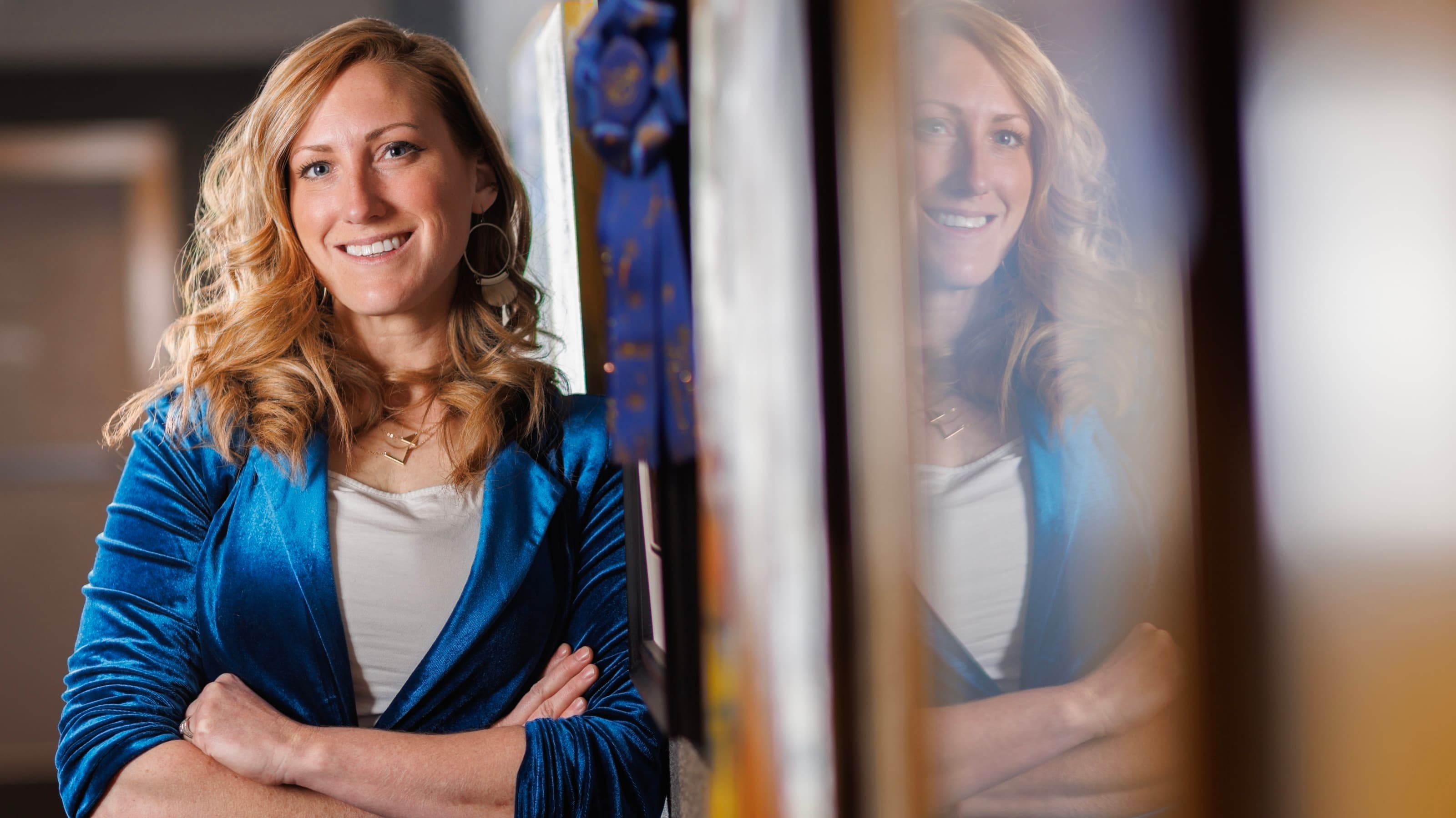 Woman in a blue blazer smiling, arms crossed, with a reflection visible in the glass nearby.
