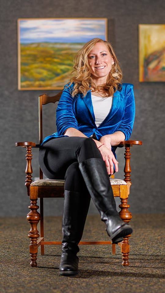 Smiling woman in a blue blazer sitting on a wooden chair, with paintings in the background.