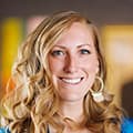 Smiling woman with curly blonde hair and large hoop earrings, colorful background blurred.