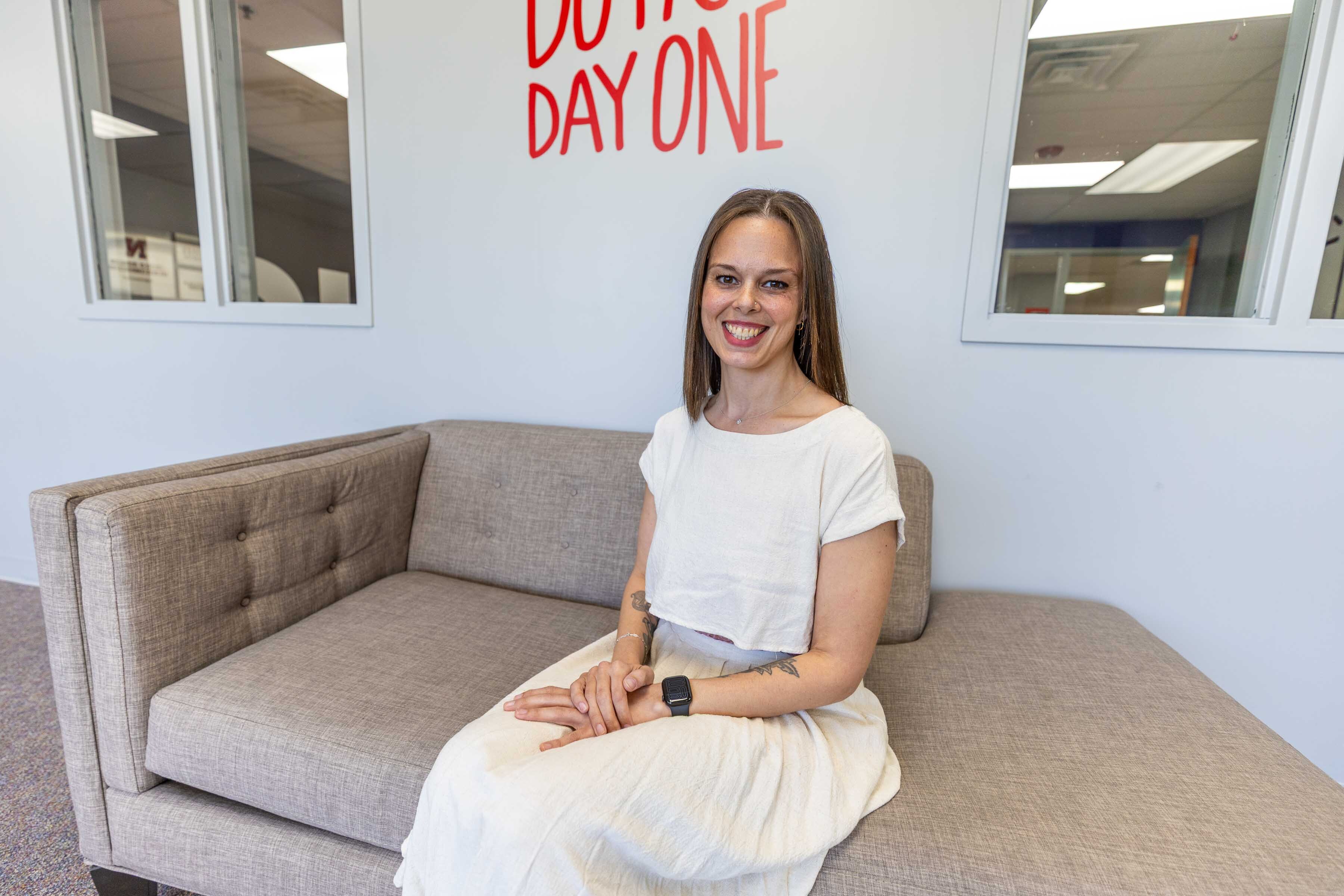 Woman seated on a gray couch in a bright office space, with a motivational phrase on the wall.