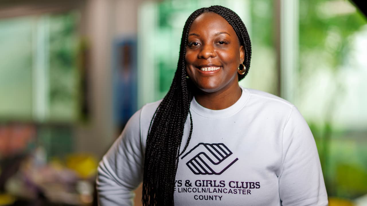 Woman smiling indoors, wearing a white Boys and Girls Clubs sweatshirt, with long braided hair.