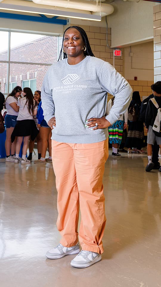 Woman smiling in a school hallway, wearing a gray sweatshirt and peach pants. Students in the background.
