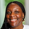 Smiling woman with braided hair, gold hoop earrings, and a white shirt, against a blurred green background.