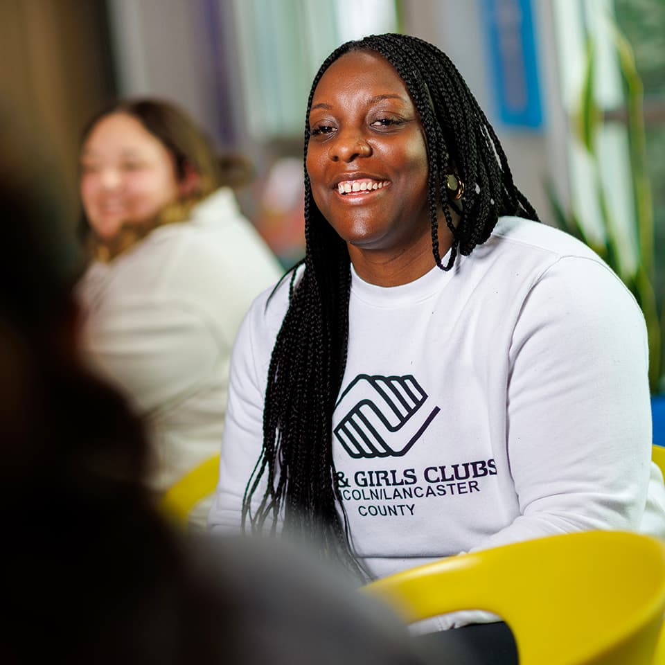 Smiling woman wearing a white Boys and Girls Clubs sweatshirt, seated in a bright space.