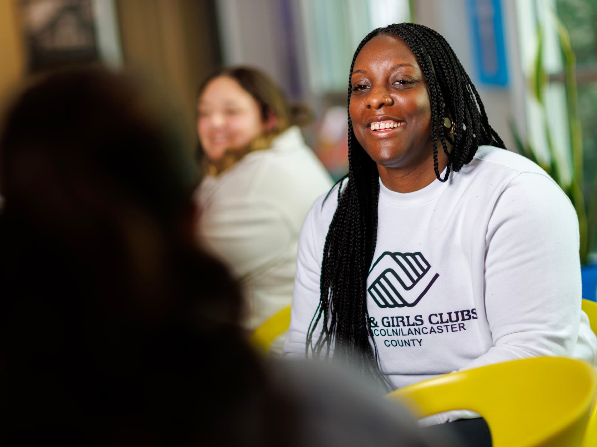 Smiling woman in a white Boys & Girls Clubs sweatshirt seated in a bright room.