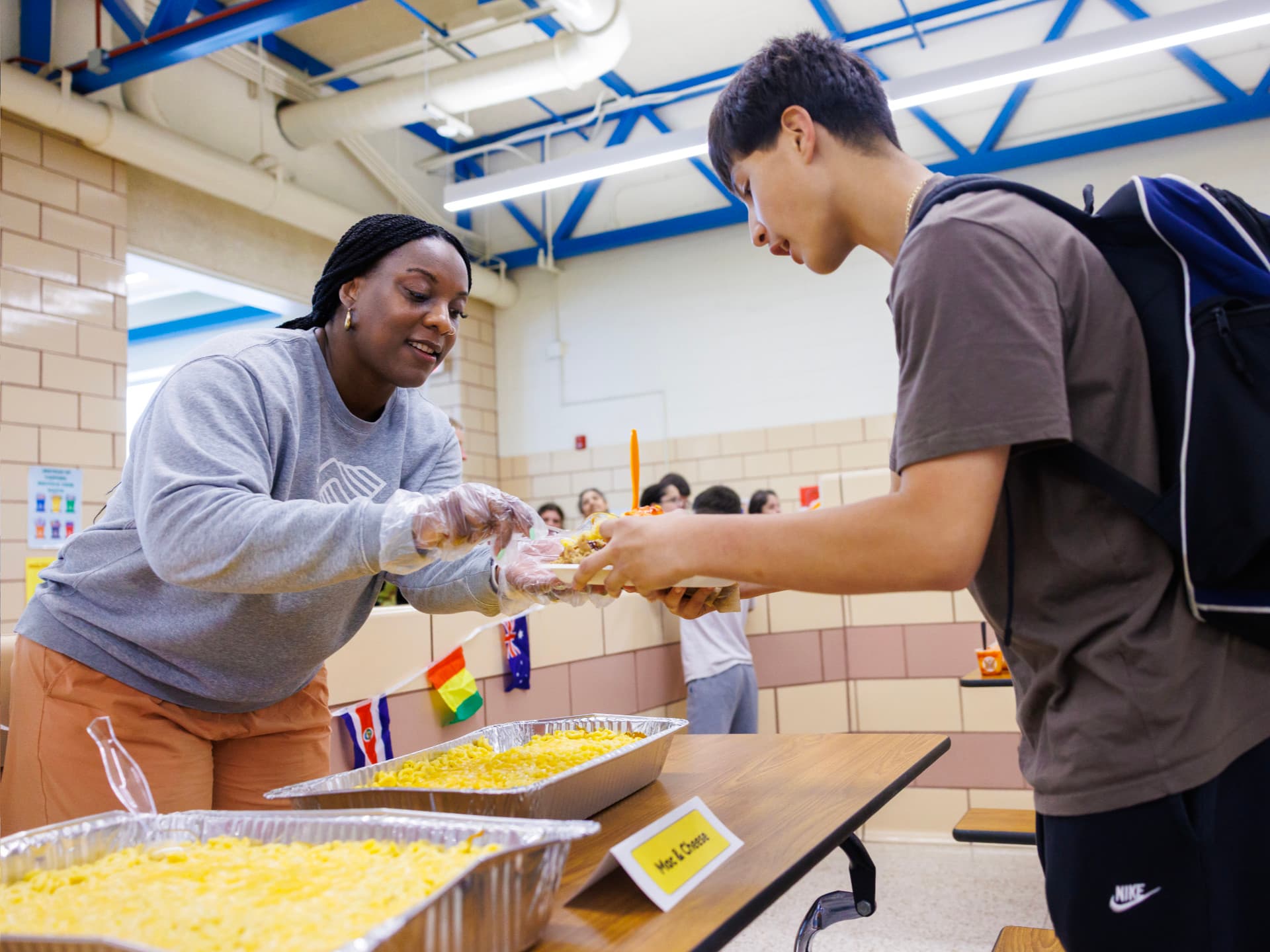 Server hands food to student in a school cafeteria, dishes labeled with flags on a table.