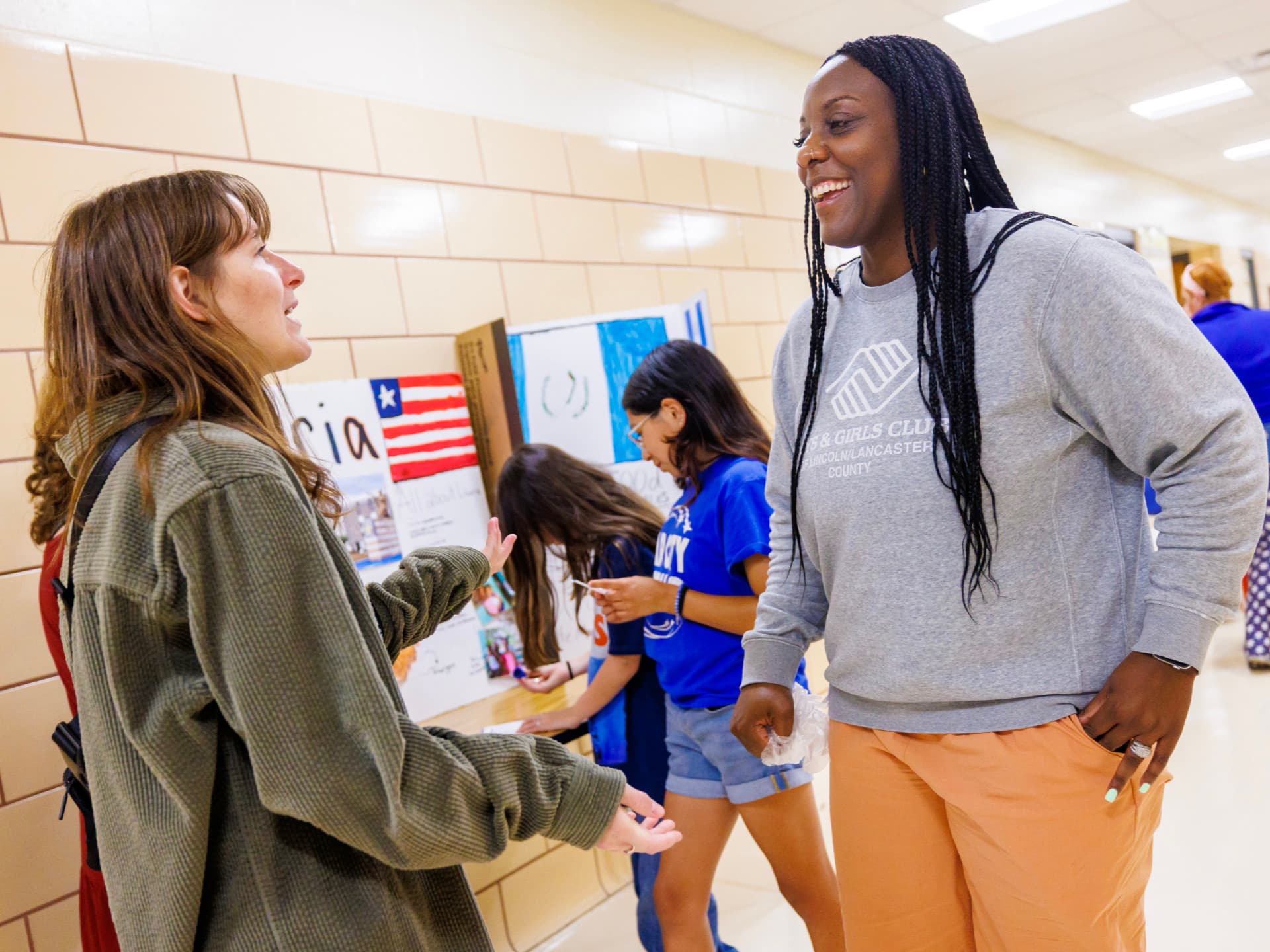 Two women converse in a school hallway near students and project displays.