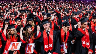 Graduates in caps and gowns with red sashes, holding their degrees, turn their tassels in a crowded ceremony.
