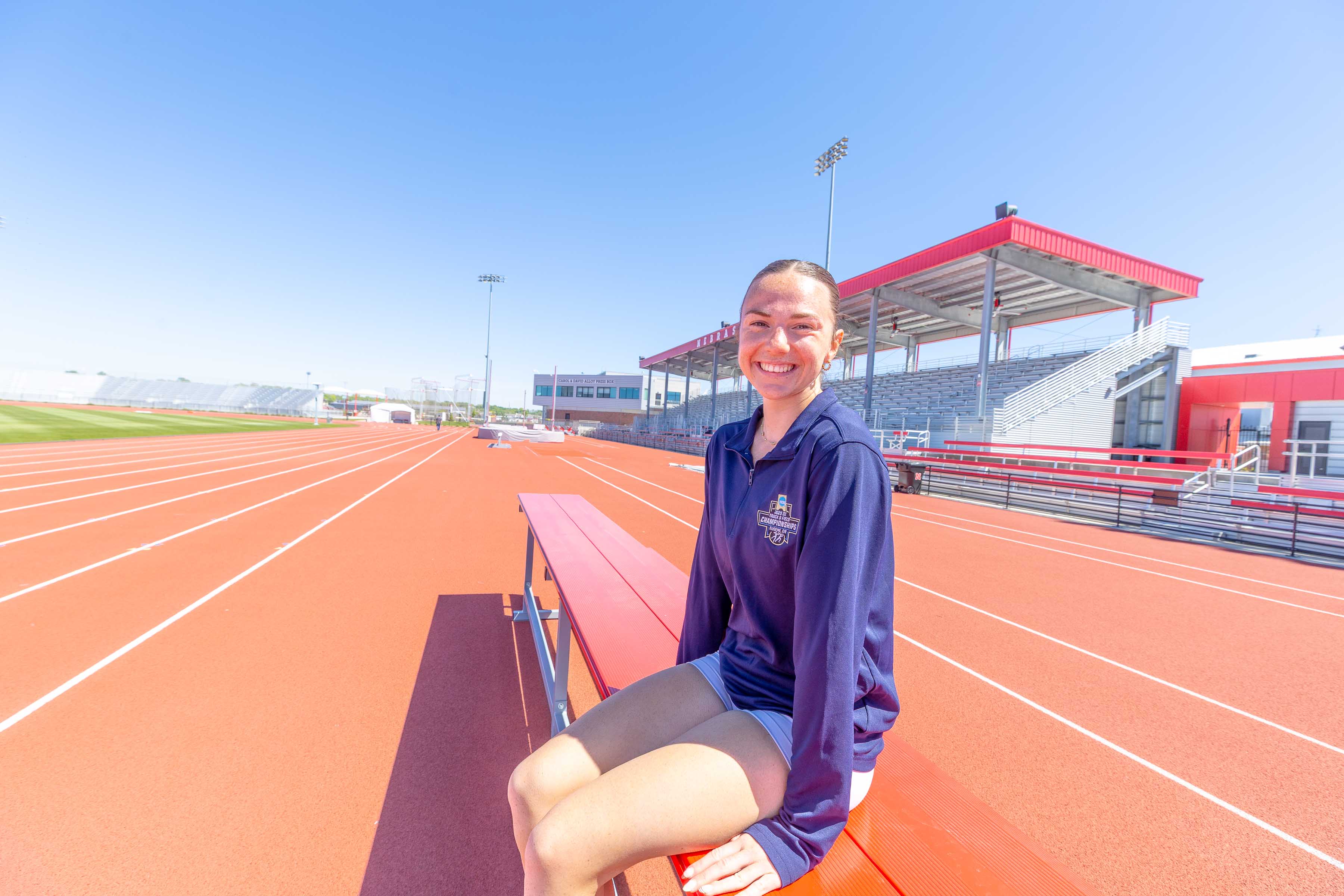 Person sitting on red bleachers at a track and field stadium under a clear blue sky.