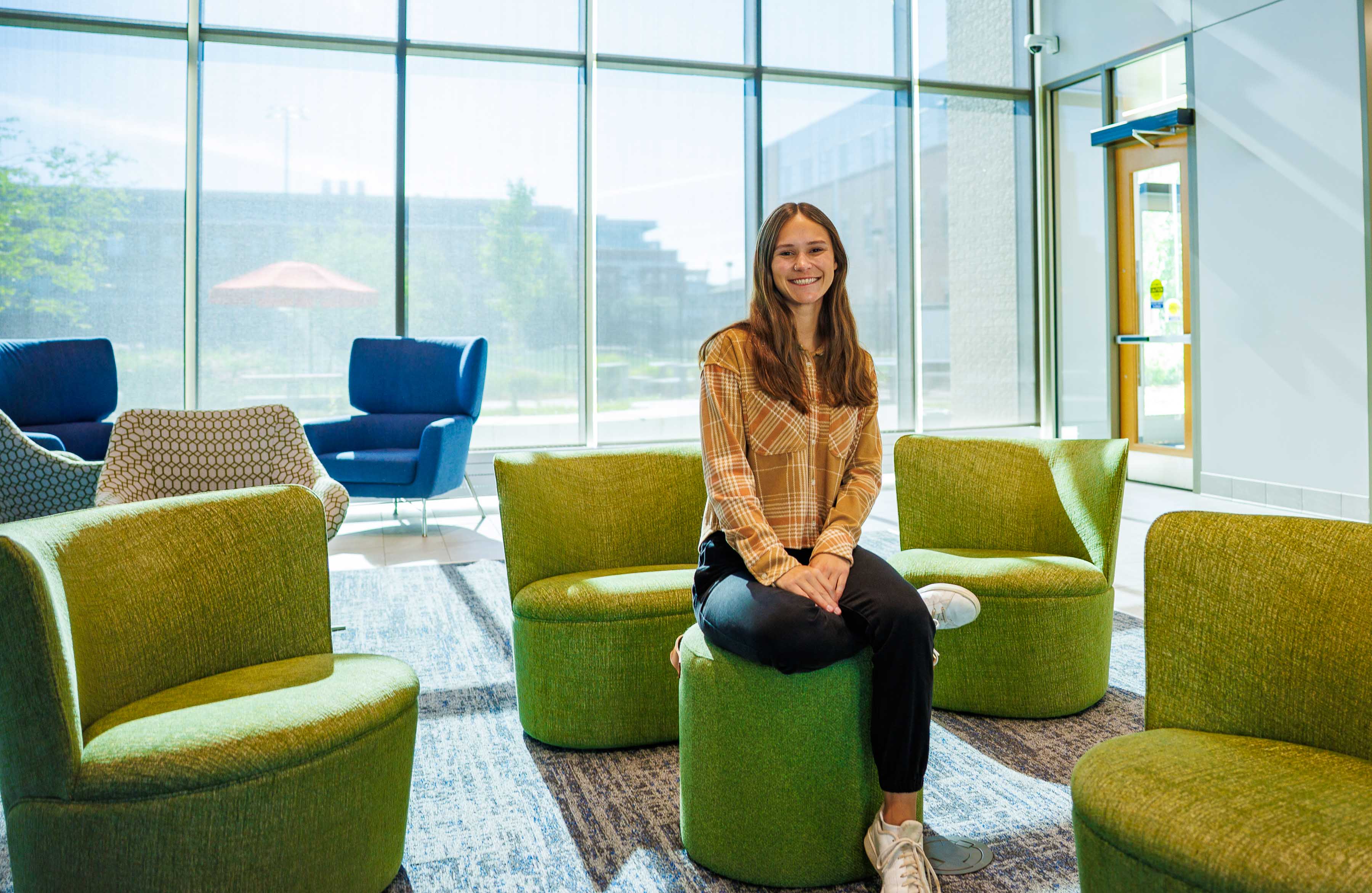Woman seated on a green ottoman in a bright modern lounge with large windows.