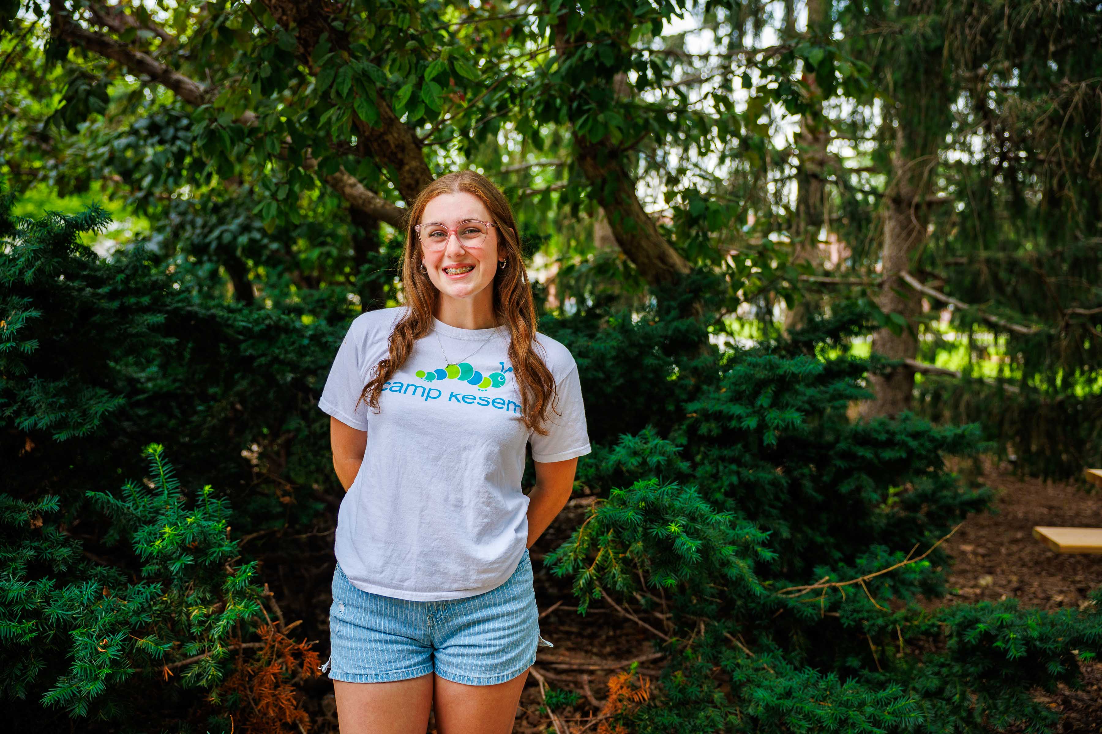A person smiling outdoors, wearing glasses, a Camp Kesem shirt, and blue shorts, surrounded by green trees.