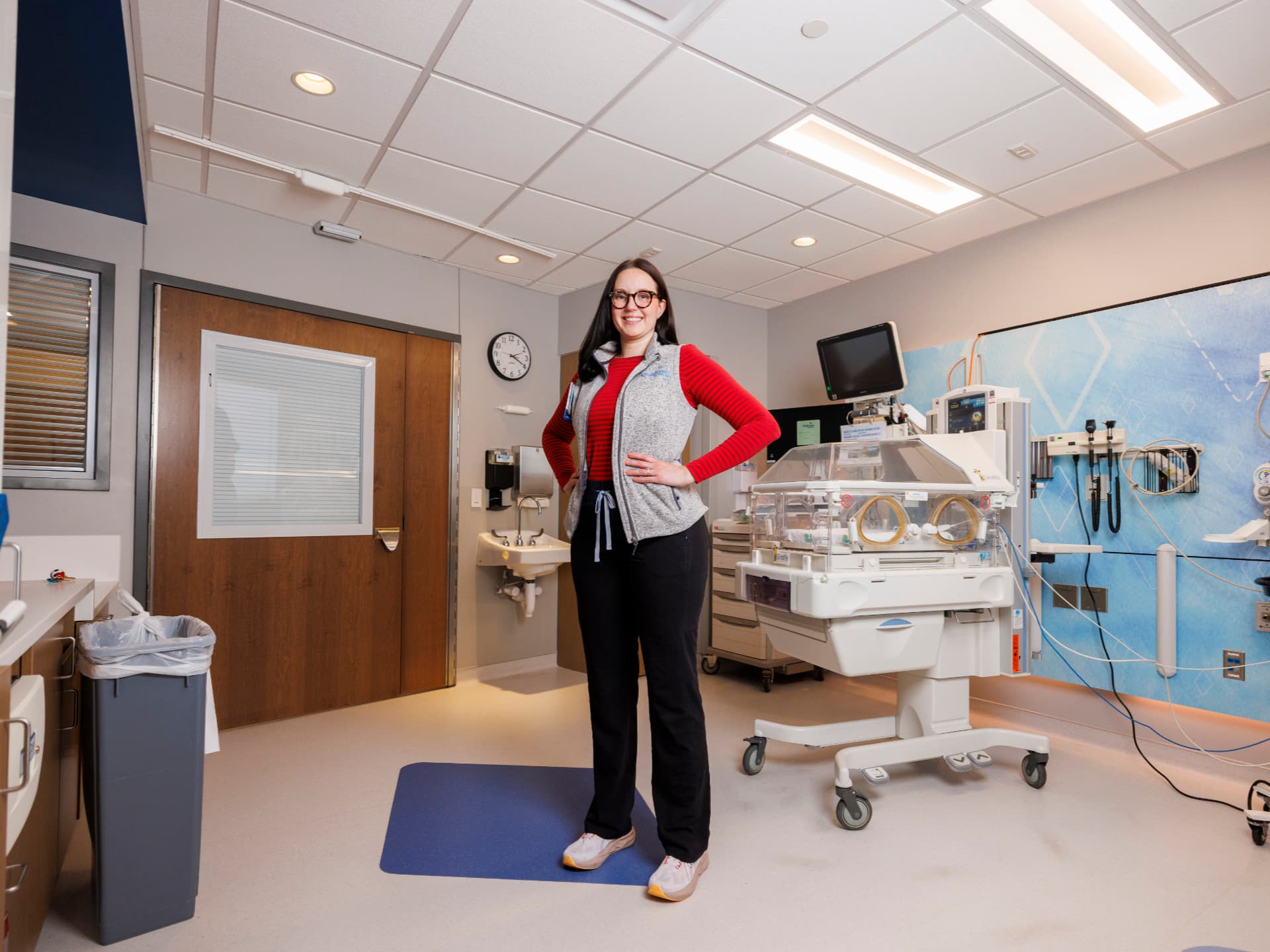 Woman standing confidently in a hospital neonatal care room with medical equipment and bright lighting.