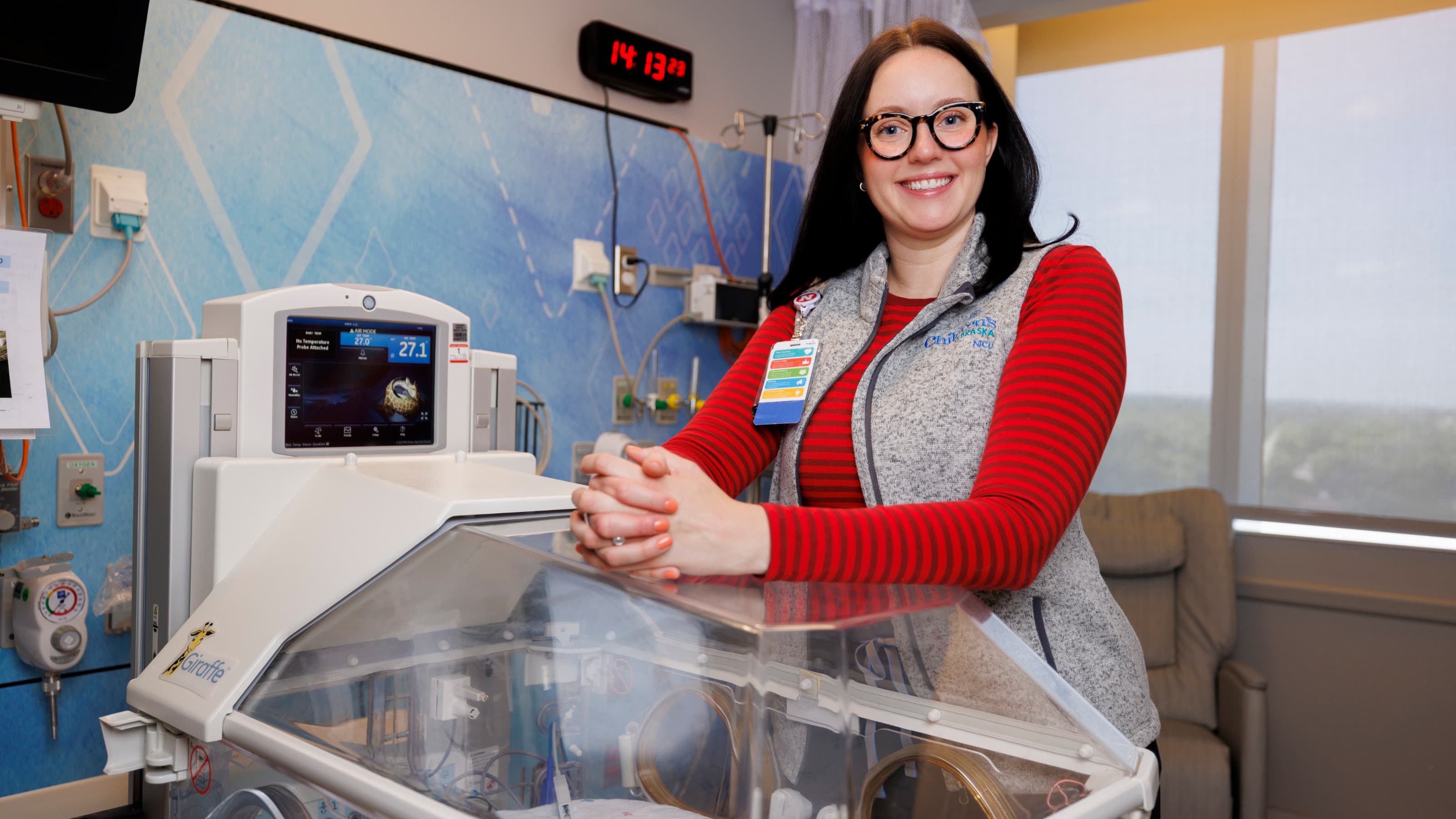 Smiling woman in glasses and red shirt standing beside a neonatal incubator in a bright hospital room.