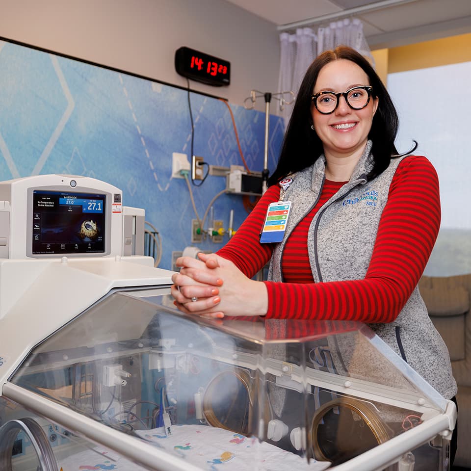 Smiling woman in a striped shirt and vest standing by a neonatal incubator in a hospital room.