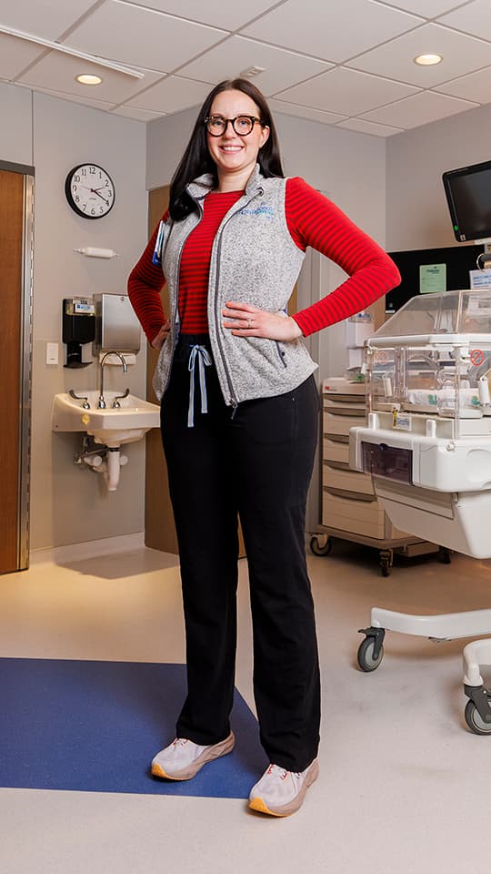 Woman stands confidently in a hospital room near an infant incubator and medical equipment.