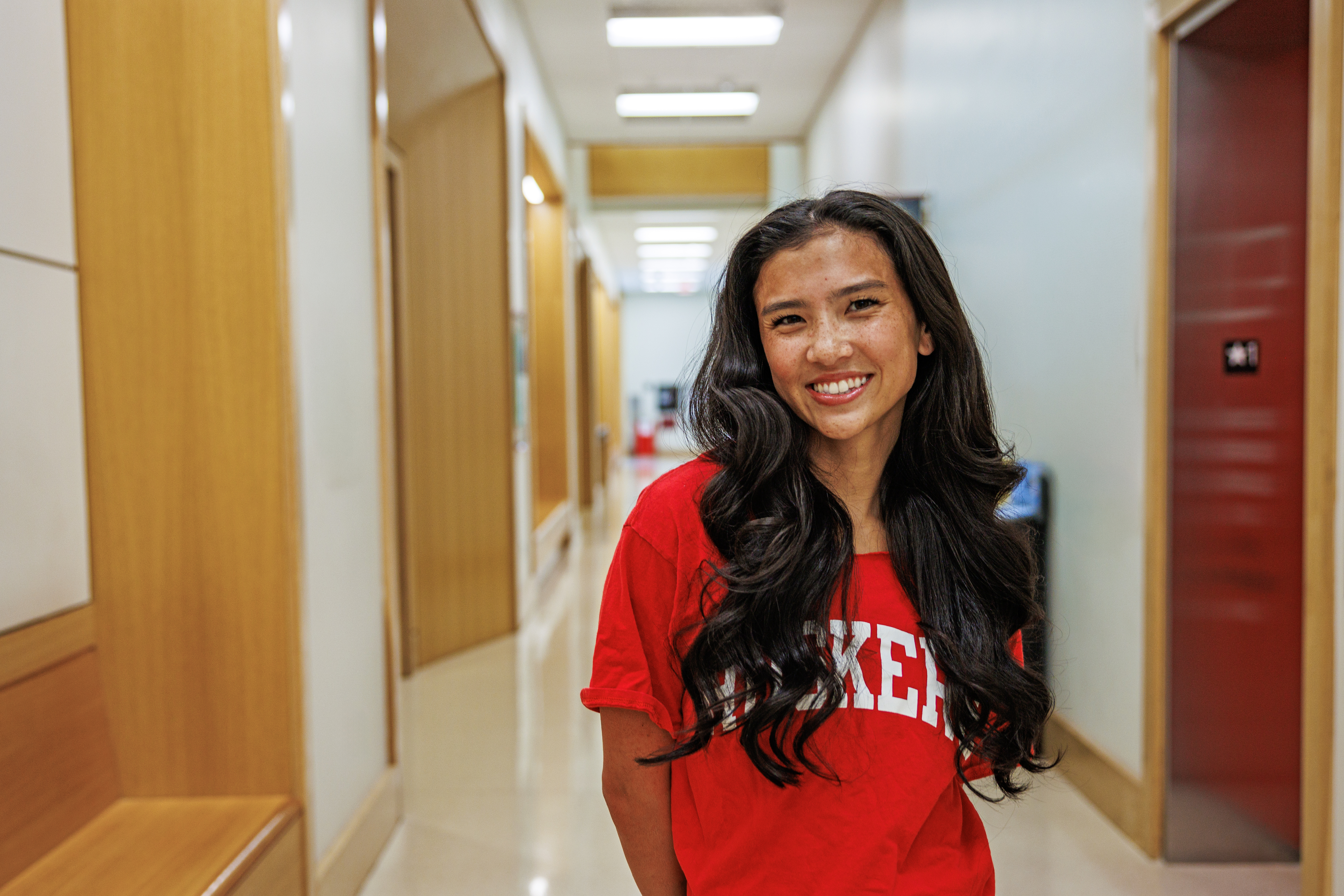 Smiling woman with long hair in a red shirt standing in a brightly lit hallway.