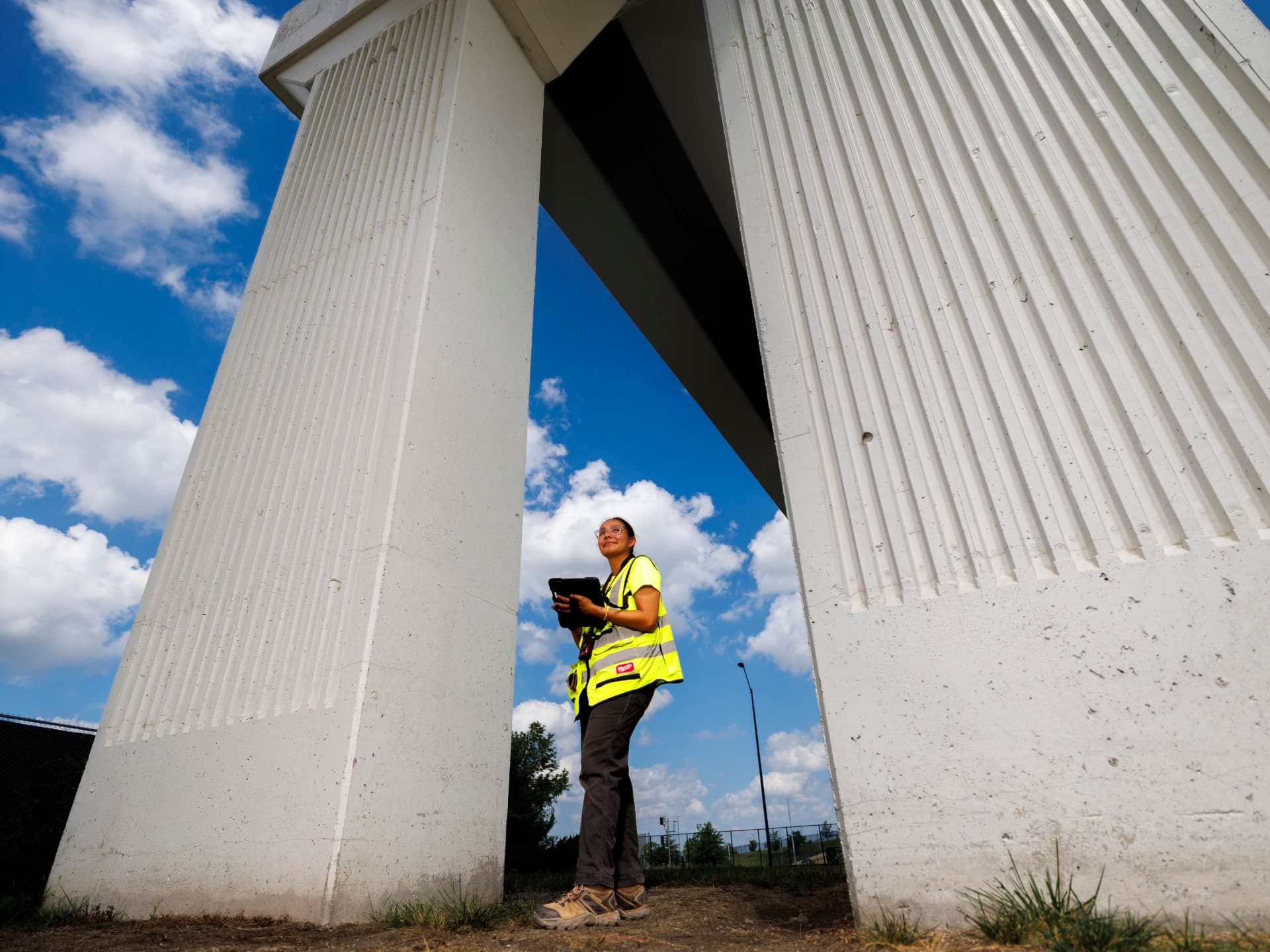 Worker in a yellow vest stands under a large white structure on a sunny day with scattered clouds.